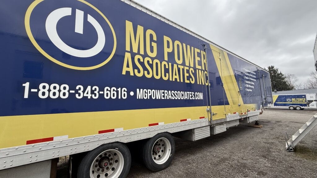 Blue and yellow MG Power Associates Inc trailer with large logo and contact number on the side, shown in a parking lot.