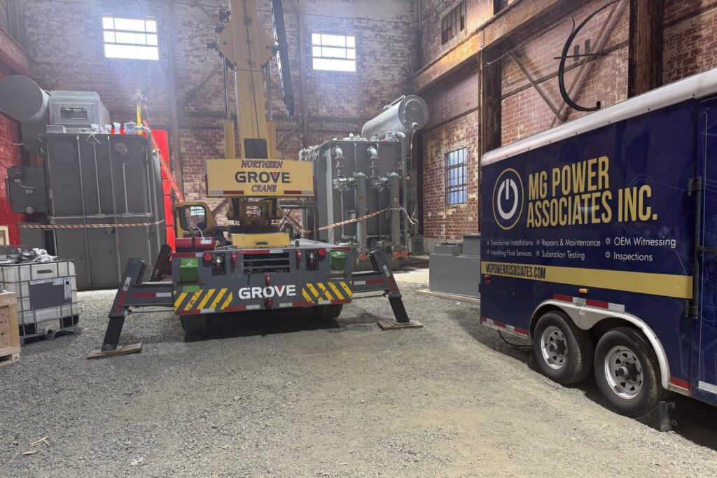 Large mobile crane on a trailer inside a brick-walled warehouse, with a blue MG Power Associates trailer nearby.