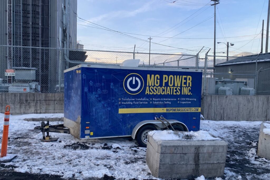 Blue MG Power Associates Inc trailer in an industrial lot, with snow on the ground and chain-link fencing in the background.